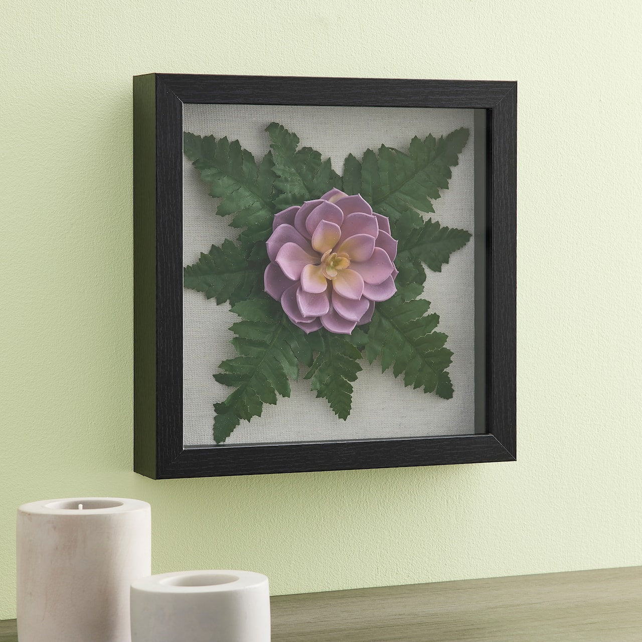 Black shadow box frame holding purple flower and green leaves on white background