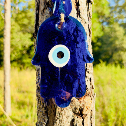 Blue hamsa hand amulet with white evil-eye circle, hanging from macrame rope